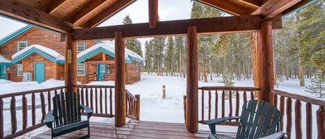 Eine Holzhütte mit überdachter Veranda, zwei Adirondack-Stühlen und verschneiter Landschaft.