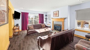 A cozy living room with a fireplace, a sofa set, a television, and a large window with curtains. - 3BR - Hot Tub, Jacuzzi, Stunning Mt. Elbert Views (Leadville)