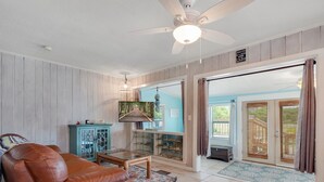 A living room with a brown leather sofa, a glass coffee table, a teal cabinet, and a television mounted on the wall. - The Beachy Pool • Cozy SGI Cottage • Pool (St. George island)