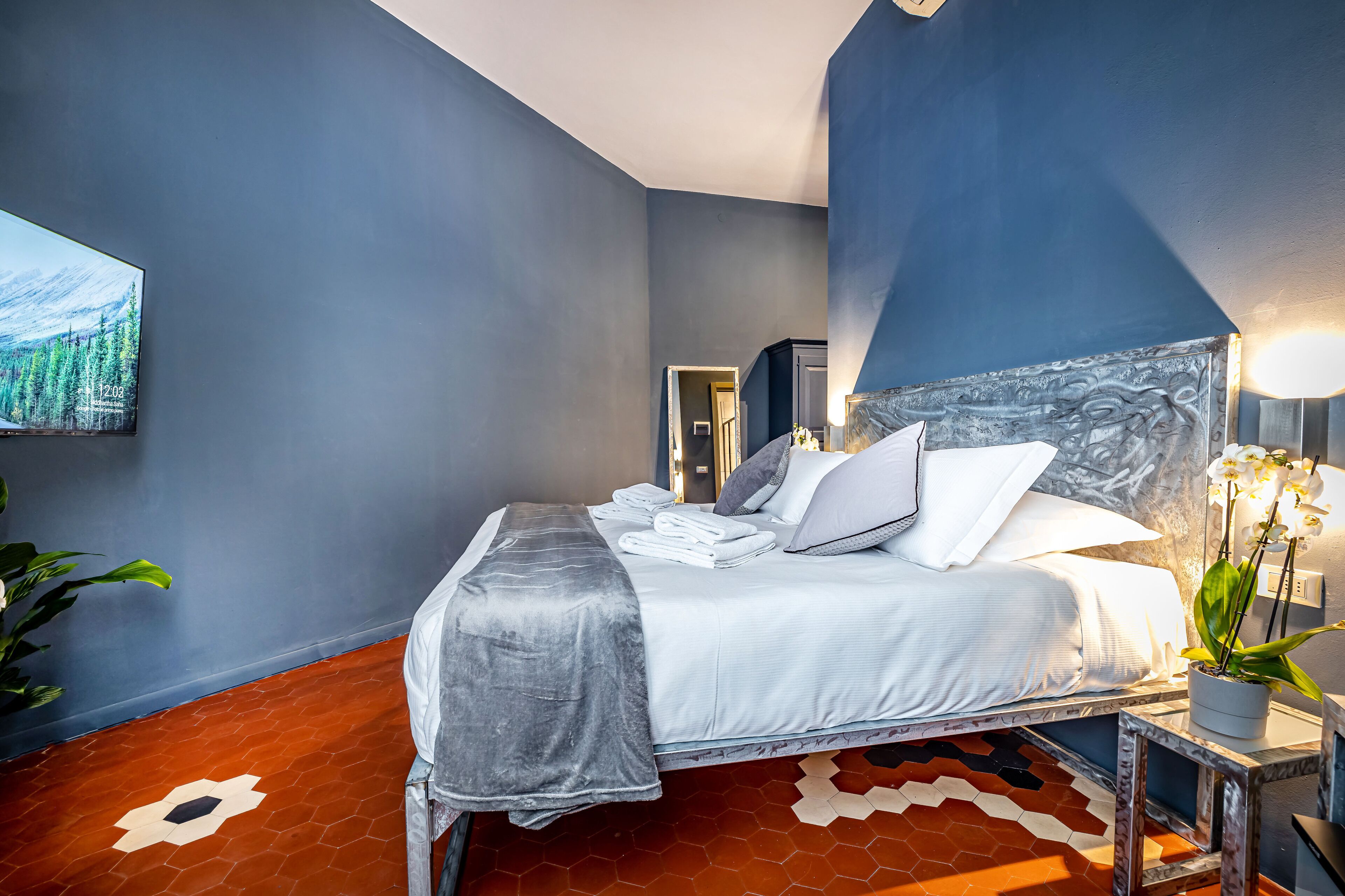 Ein ordentlich bezogenes Bett mit Kissen und Decke, ein Blumentopf auf einem Nachttisch und ein großes Bild an der Wand.