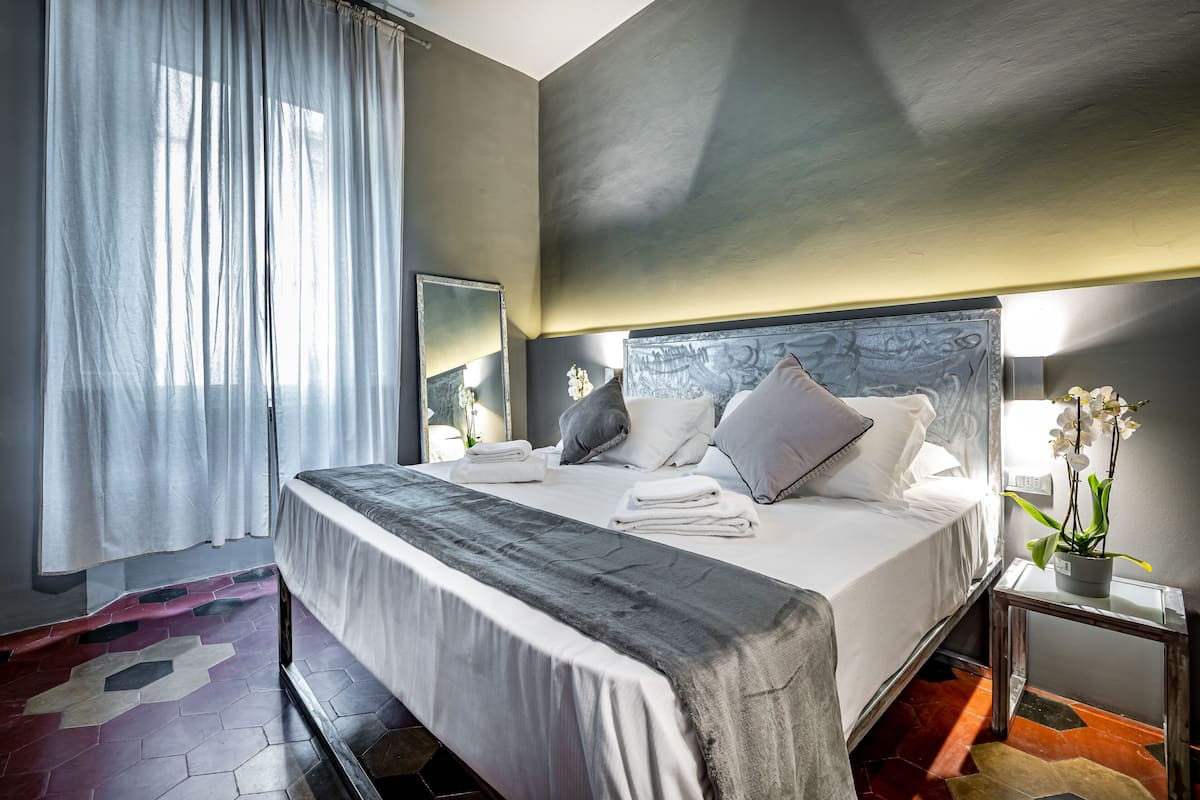 A neatly made bed with pillows and a blanket, a bedside table with a plant, a mirror, and a window with curtains.