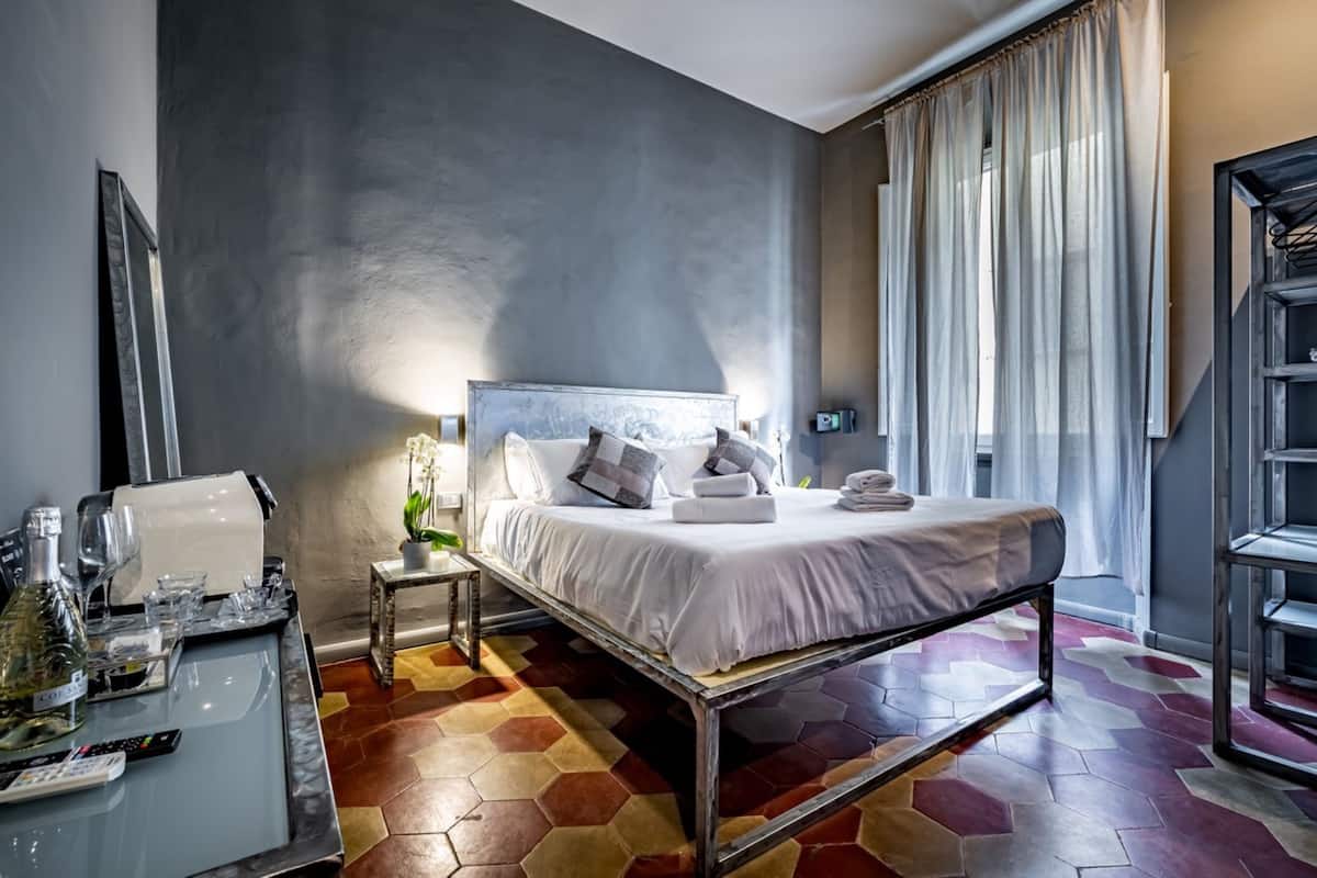 A modern hotel room with a large bed, a bedside table with a lamp, a small side table, and a glass-topped table with a bottle and glasses.