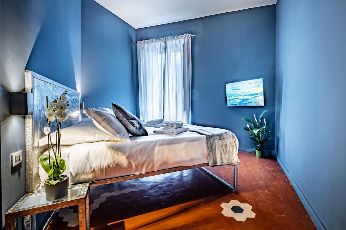 A bedroom with a bed, a potted plant, a painting, and a window with curtains.