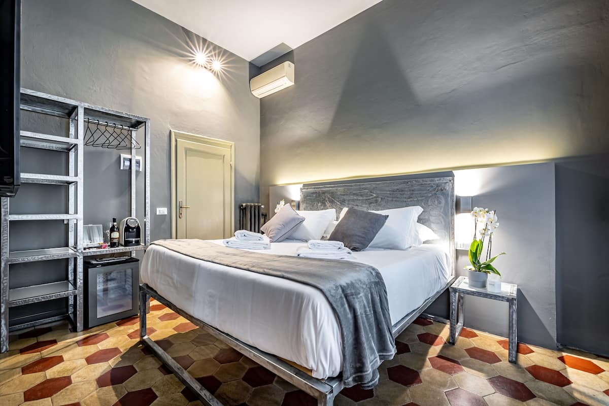 A hotel room with a bed, a small table, a shelf unit, and a radiator.