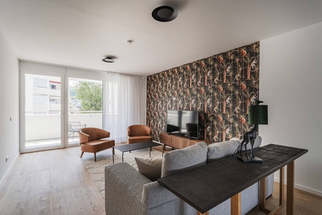 Ein modernes Wohnzimmer mit Sofa, Couchtisch und Fernseher. Eine Schiebetür aus Glas führt zu einem Außenbalkon.