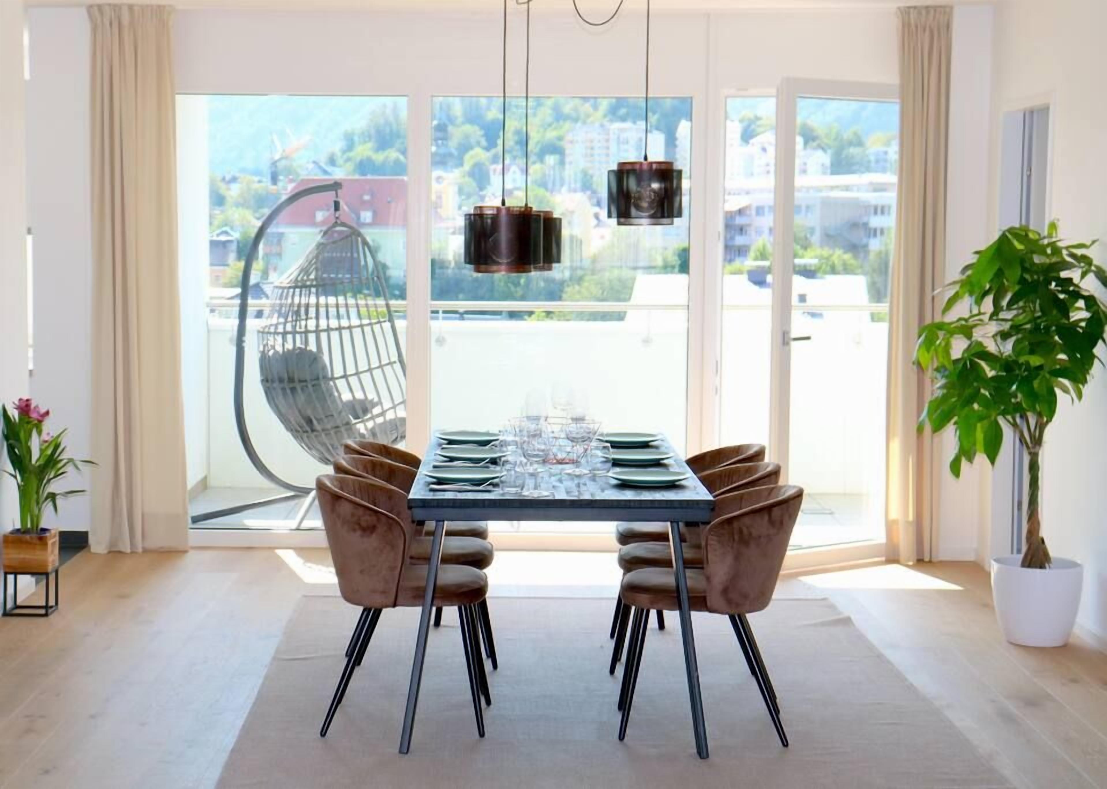 Uma área de jantar com uma mesa posta para quatro pessoas, duas cadeiras de cada lado, um lustre pendurado e uma janela grande com vista para edifícios e vegetação.