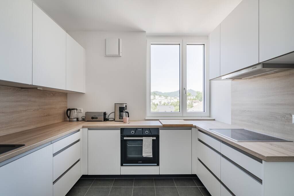 A modern kitchen with white cabinets, a built-in oven, a coffee maker, and a window with a view of the city.