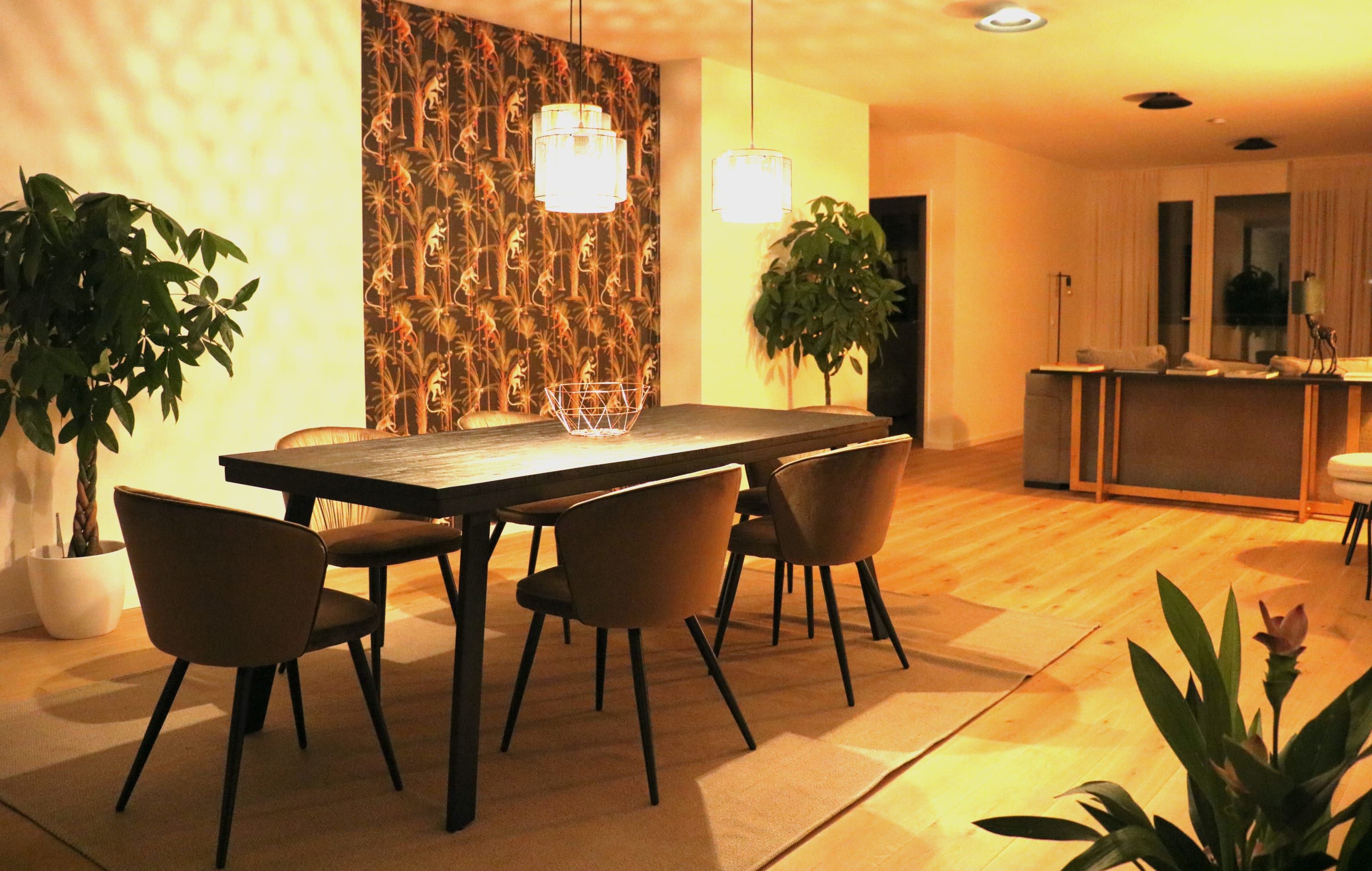 A modern dining area with a wooden table, four chairs, and two pendant lights. There are potted plants on either side of the table.