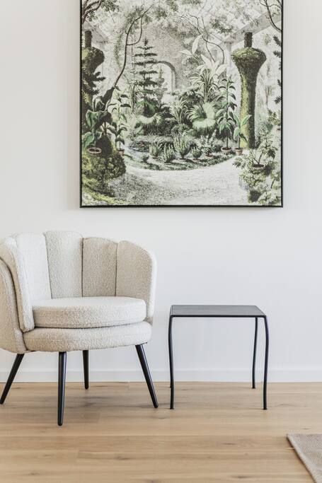 A room with a framed botanical print on the wall, a beige armchair, and a small black table.