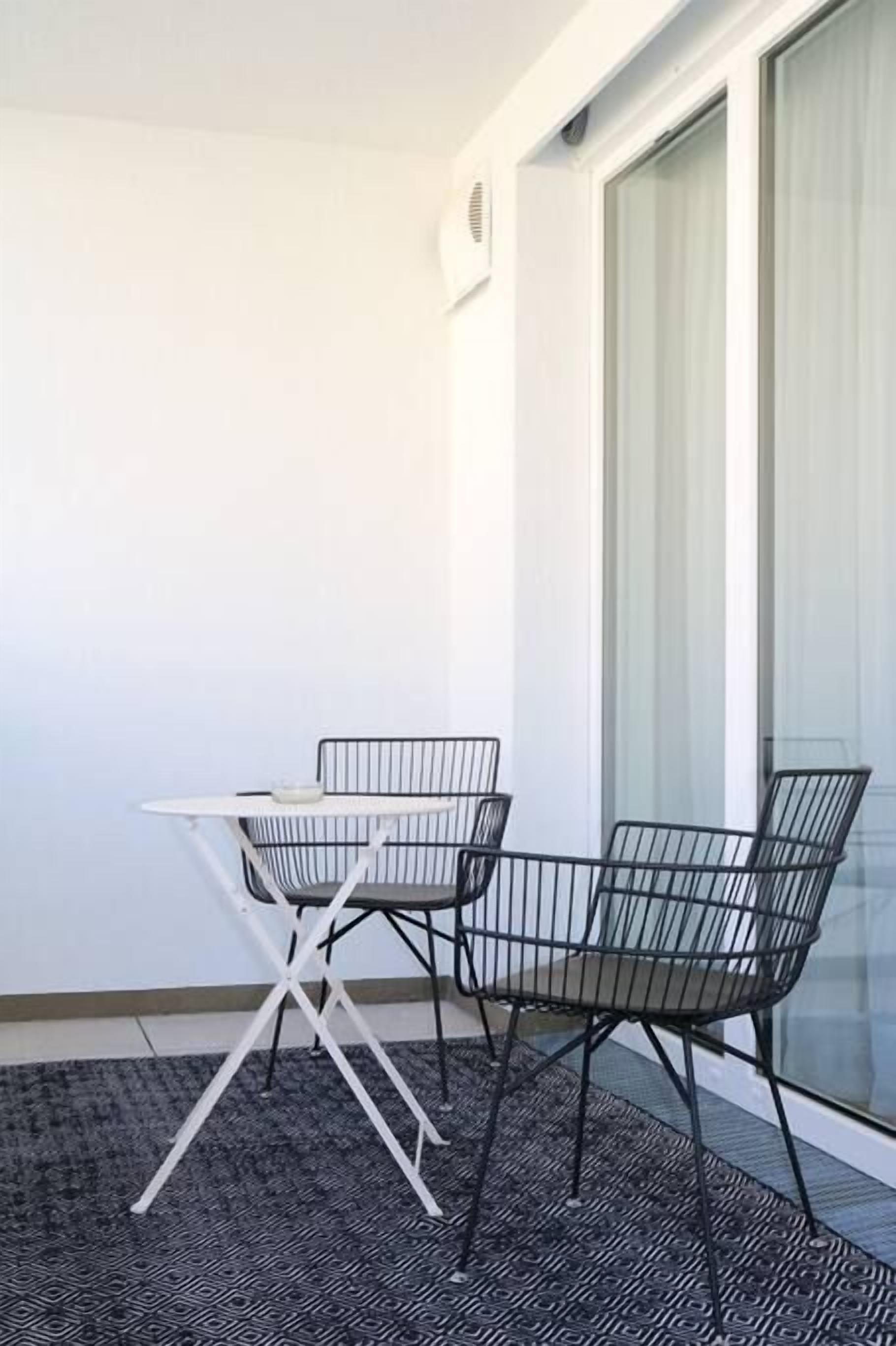 A balcony with a small table and two chairs, a patterned rug, and a window with curtains.