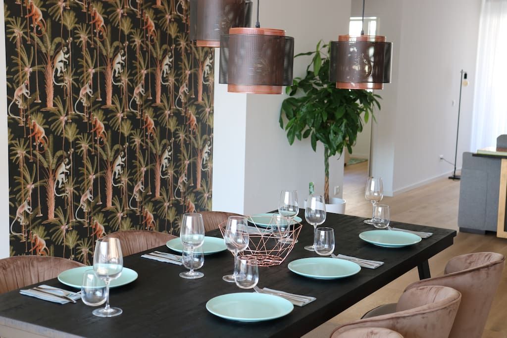 Ein Essbereich mit einem schwarzen Tisch, gedeckt für sechs Personen, hellblauen Tellern und Weingläsern. Über dem Tisch hängen Kupfer-Pendelleuchten.