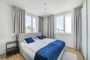 A modern bedroom with a large bed, two bedside tables, a nightstand, and a view of the city through large windows. - 3 Zimmer Apartment mit Blick auf die Stadt (Gmunden)