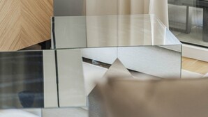 Une chambre moderne avec un téléviseur posé sur un meuble en bois, une lampe de bureau noire, une table en verre et une vue par une fenêtre avec des rideaux.
