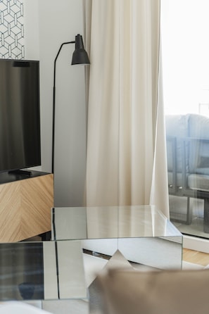Ein modernes Zimmer mit Fernseher auf einem Holzständer, einer schwarzen Schreibtischlampe, einem Glastisch und Blick durch ein mit Vorhängen drapiertes Fenster.