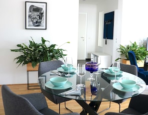 A dining area with a glass table, teal bowls, and wine glasses. There are chairs around the table, and a potted plant is visible in the background. - 3 Zimmer Apartment mit Blick auf die Stadt (Gmunden)