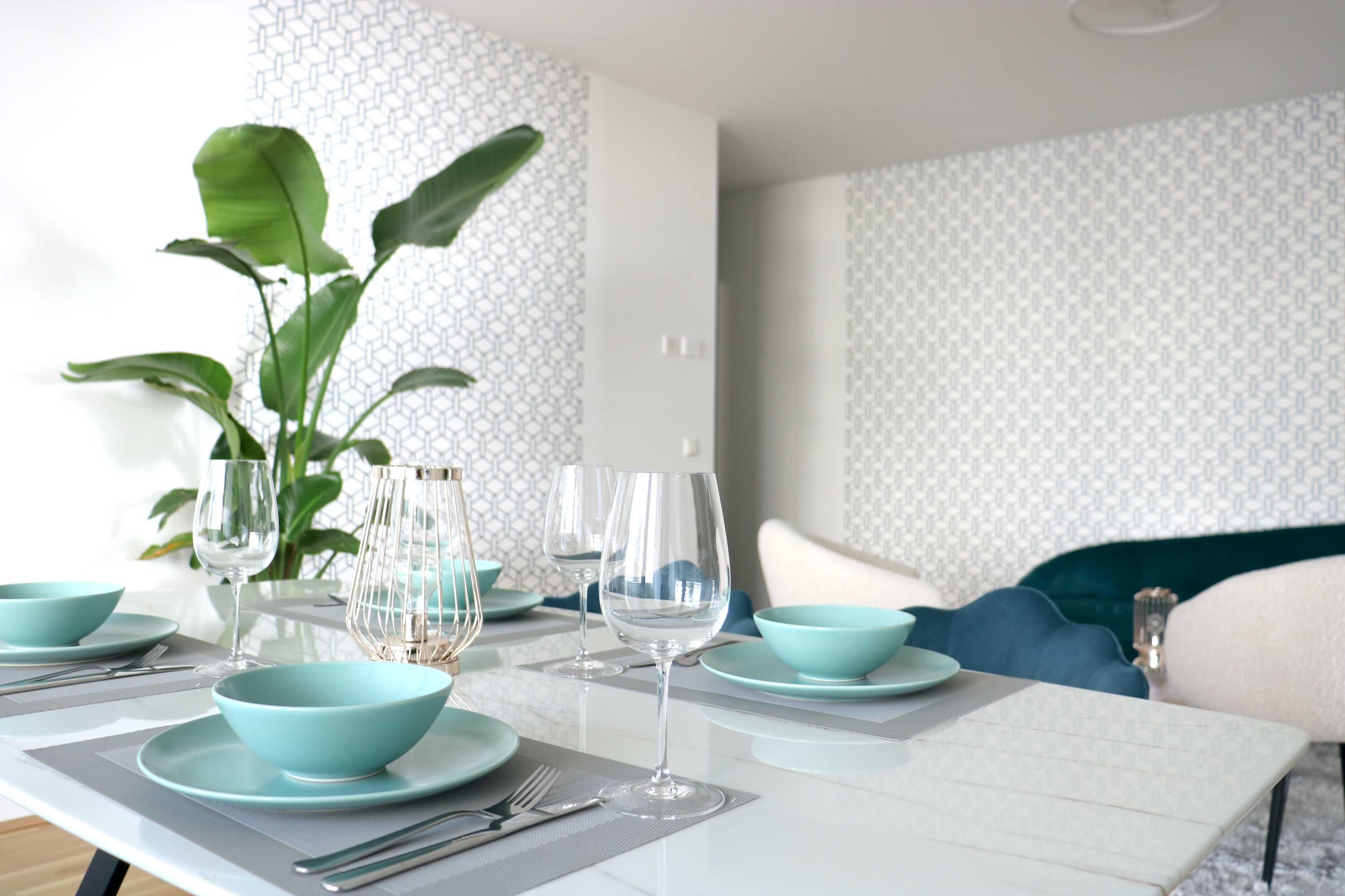 Uma mesa de jantar posta com tigelas azul-turquesa, copos de vinho e talheres, com uma planta e papel de parede estampado ao fundo.