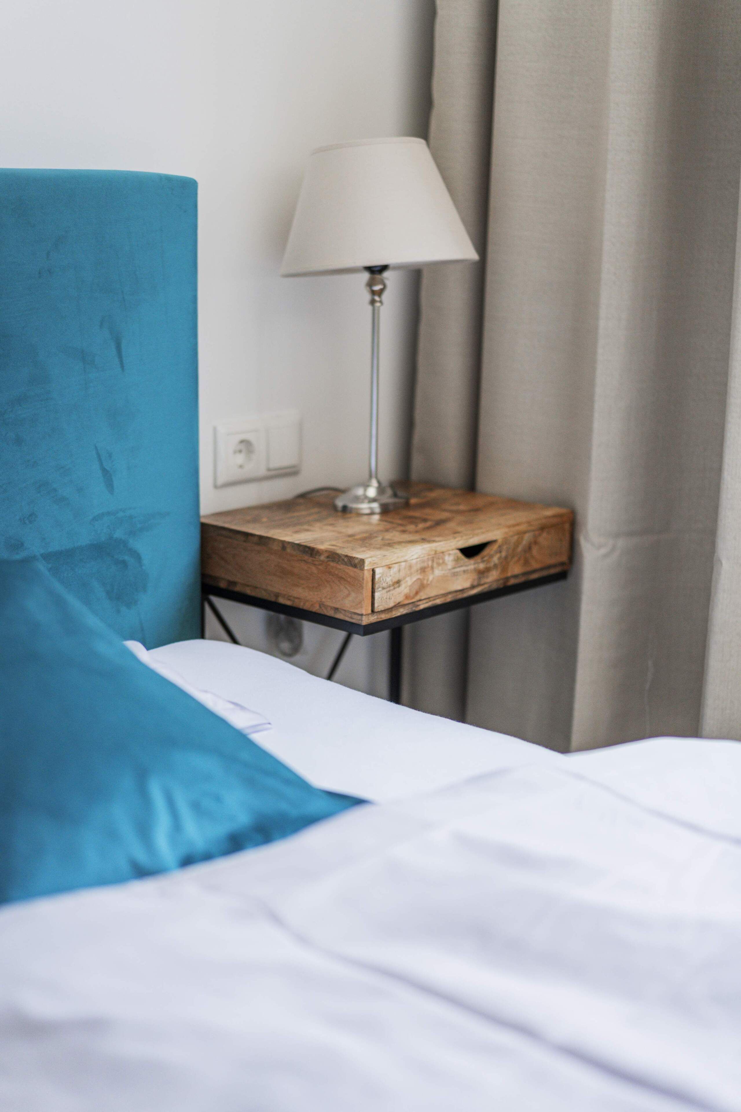 Una cama con un cojín azul, una mesita de noche con una lámpara y una cortina.
