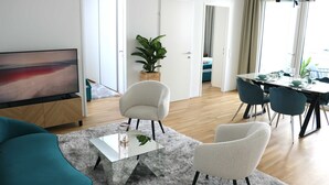 Een moderne woonkamer met een flatscreen-tv, een marineblauwe bank, witte fauteuils, een glazen salontafel en een eethoek met een tafel gedekt voor vier personen.