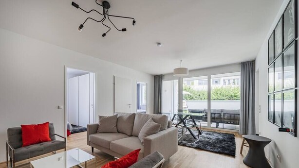 Sala de estar moderna com sofá, poltronas e mesa de centro. Há também um espaço de jantar com mesa e cadeiras. Janelas amplas oferecem vista para o exterior.