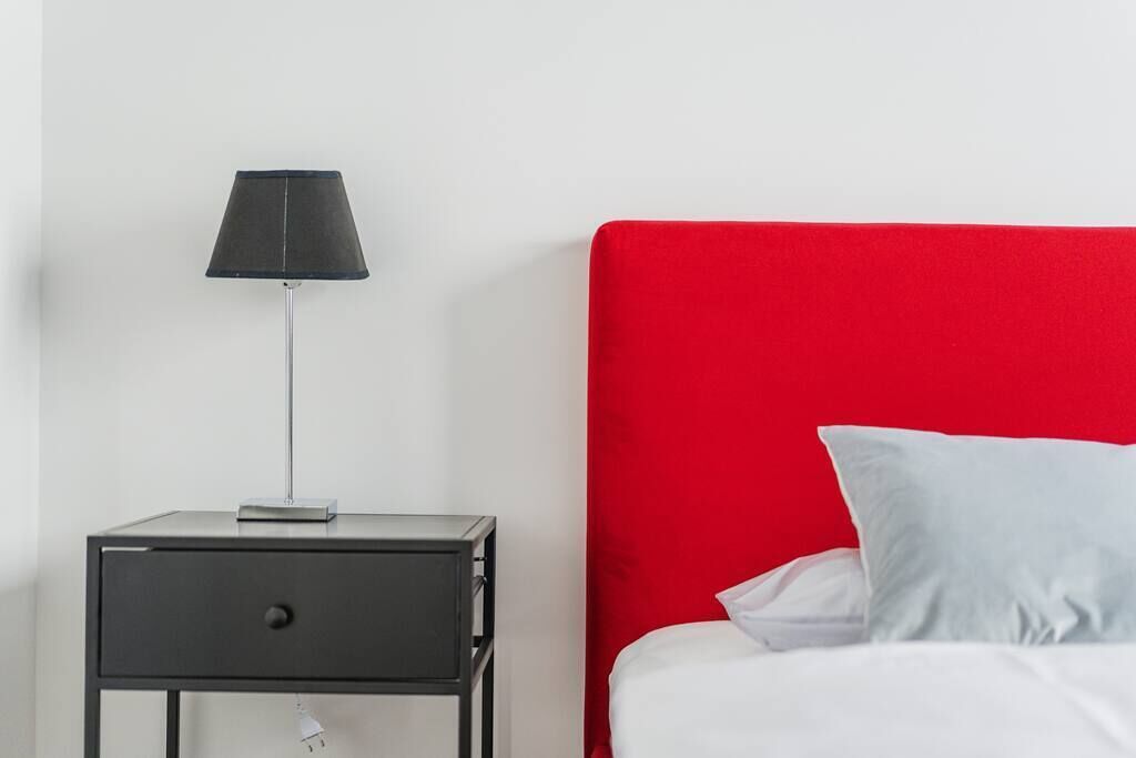 Un letto con testiera rossa e biancheria bianca, un comodino con lampada e una presa elettrica sulla parete.