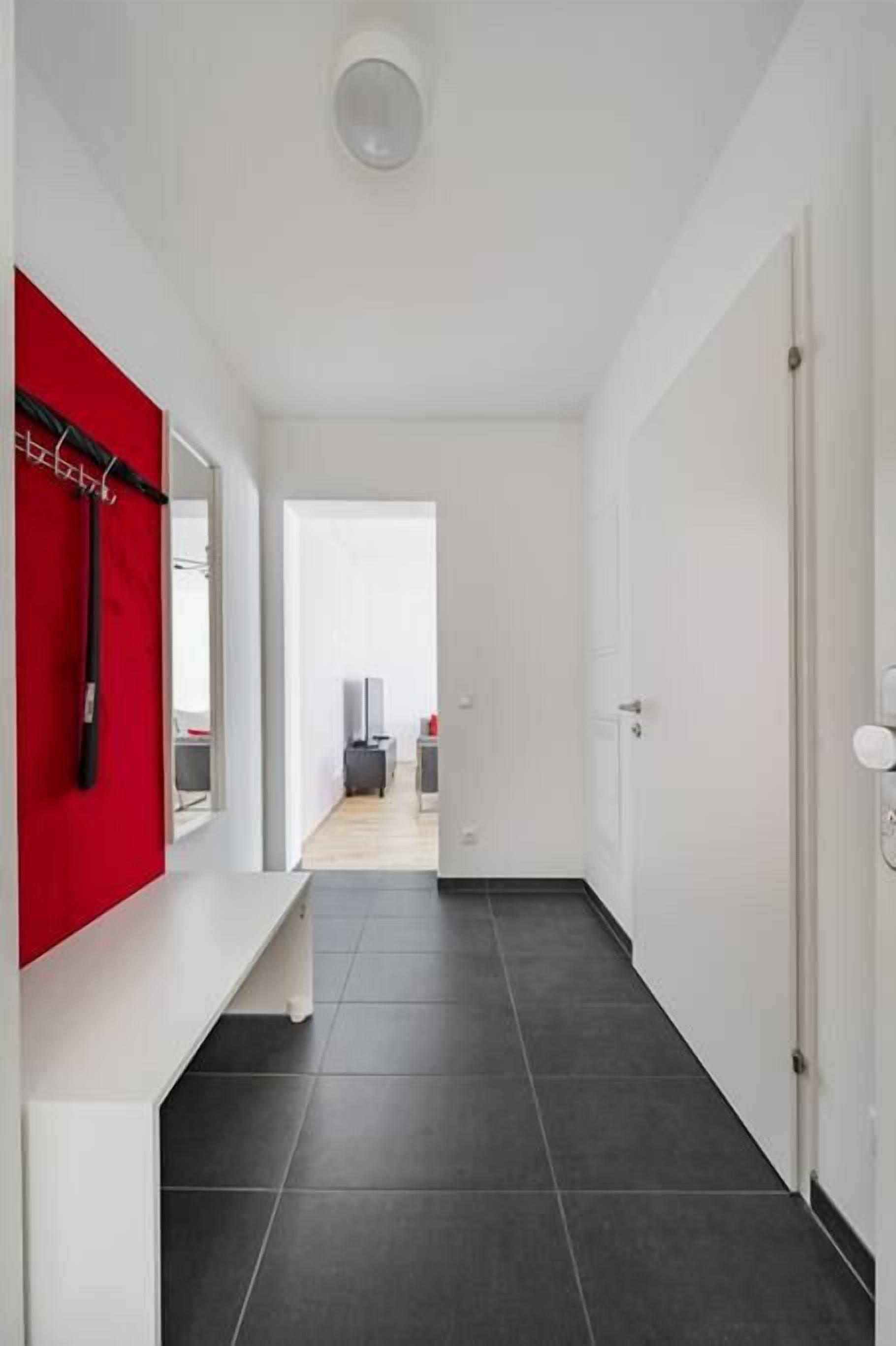 Un pasillo moderno con una pared de acento roja, una puerta blanca y vistas a una habitación con cama y televisión.