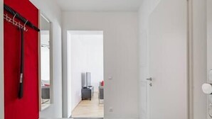 Um corredor moderno com uma parede de destaque vermelha, uma porta branca e vista para um quarto com cama e televisão.