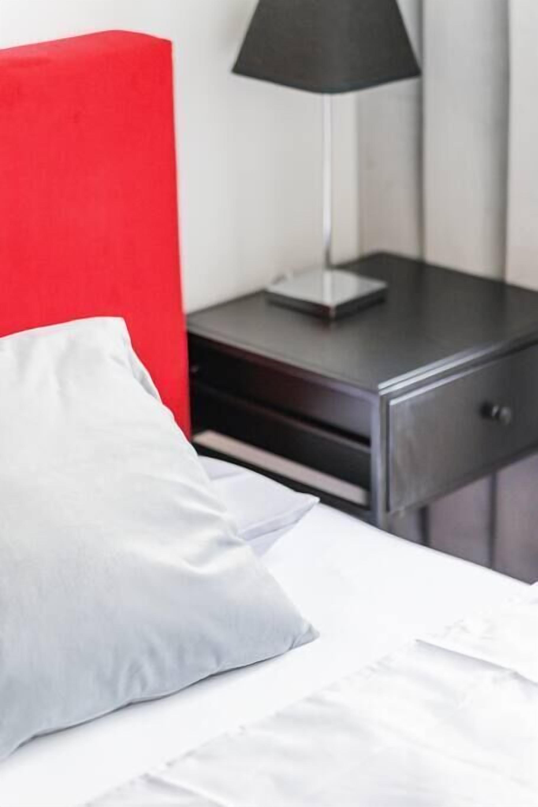 A bed with white bedding and a grey pillow, a black lamp on a bedside table, and a red headboard.