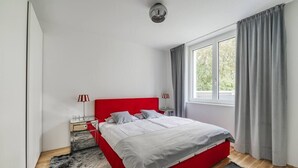 Quarto moderno com cama vermelha, mesa de cabeceira pequena, luminária e janela com cortinas.