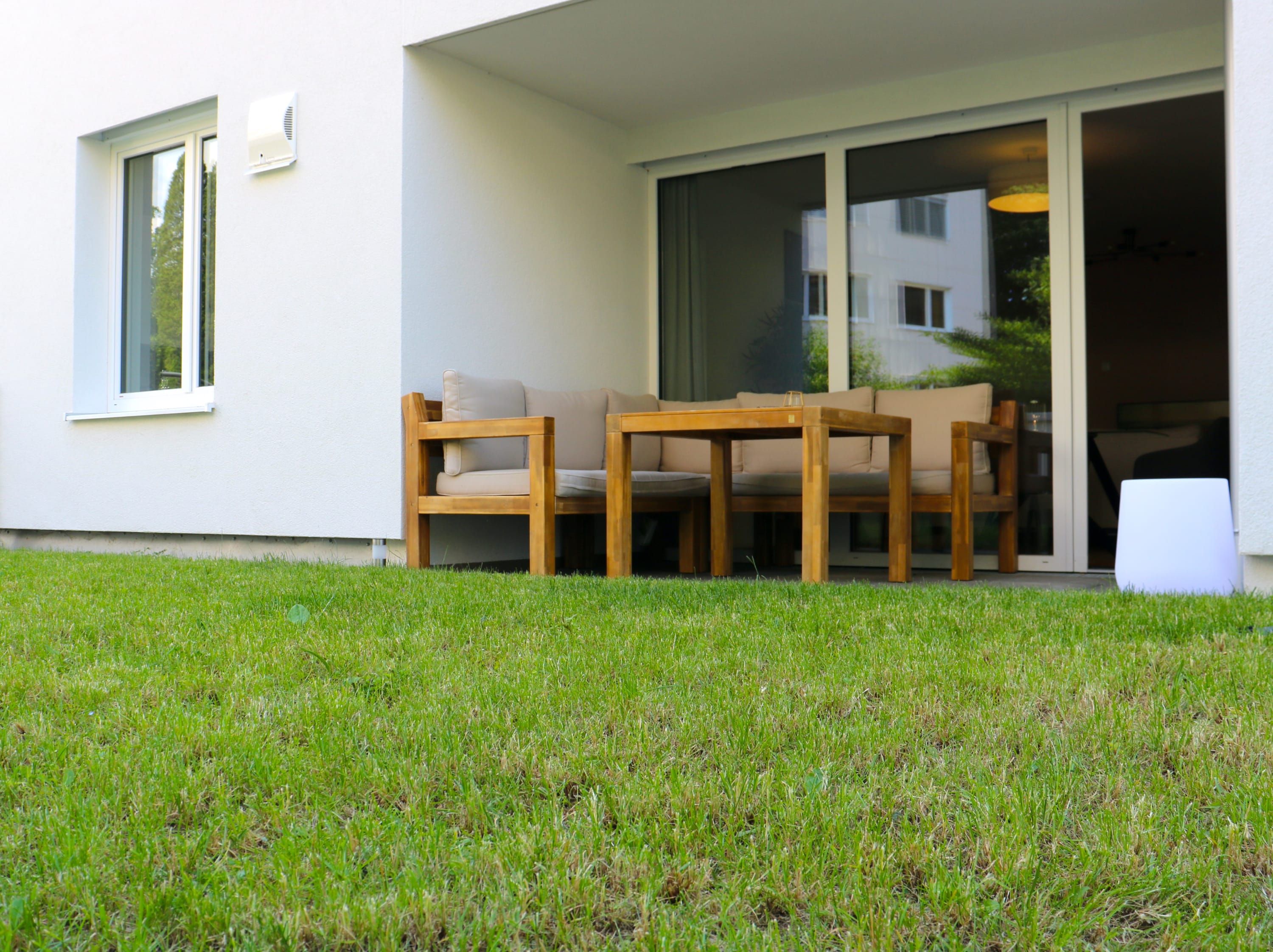 A modern outdoor seating area with a table and chairs, a grassy lawn, and a building entrance with a sliding glass door.