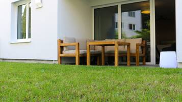 A modern outdoor seating area with a table and chairs, a grassy lawn, and a building entrance with a sliding glass door.