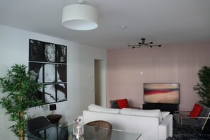 Een moderne woonkamer met een glazen salontafel, een witte bank met rode kussens, een flatscreen televisie en een groot abstract schilderij aan de muur.