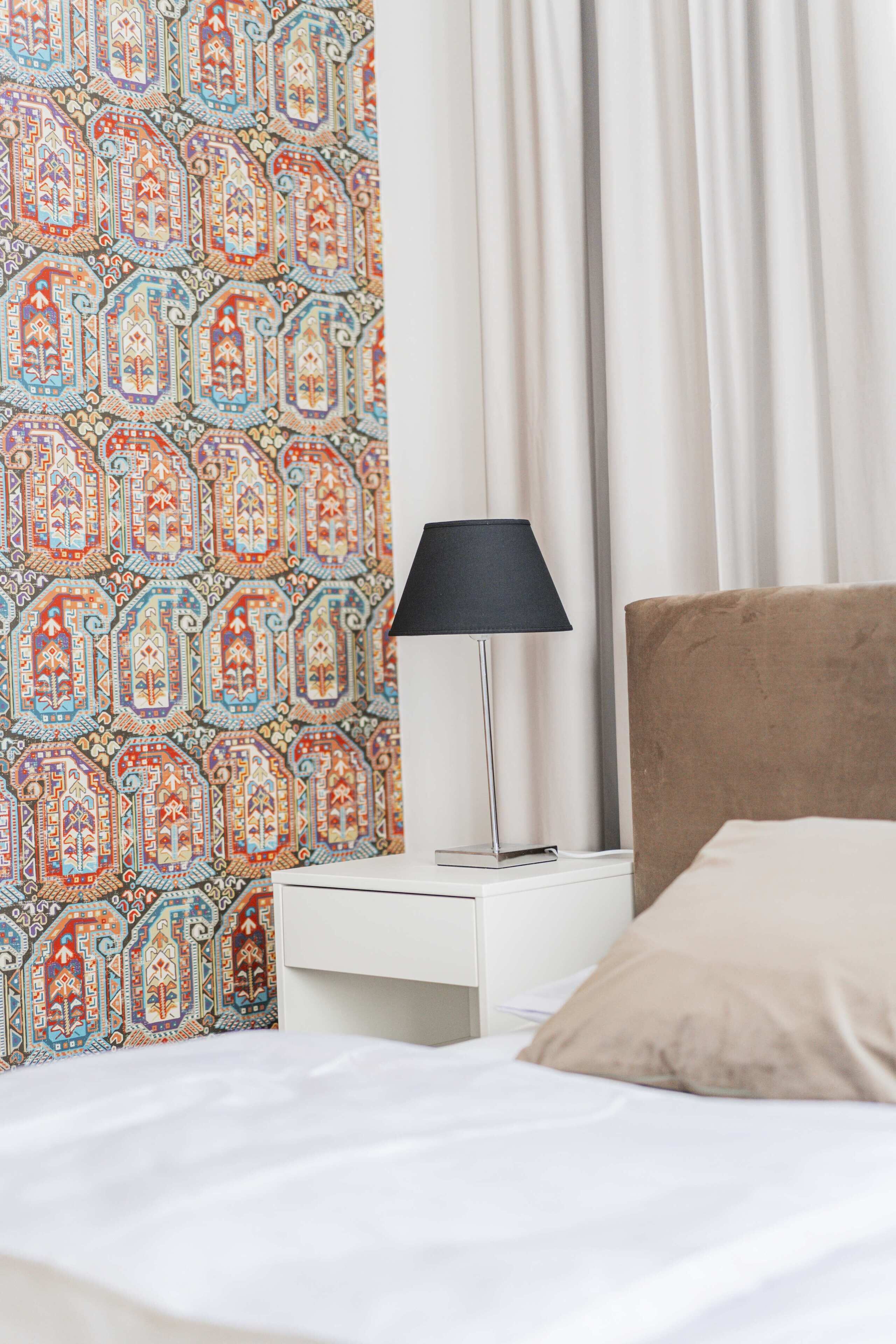 Una cama bien hecha con un almohilar beige, una mesita de noche blanca con una lámpara negra y una pared con estampado.