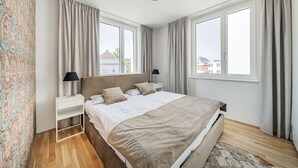 A bedroom with a large bed, two bedside tables, a nightstand, a ceiling light, and a view of buildings through the windows. - 3 Zimmer Apartment mit Blick auf die Stadt (Gmunden)