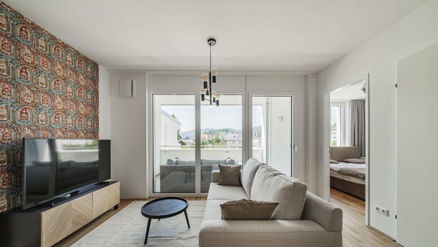 A modern living room with a flat-screen TV, a sofa, a small table, and a patterned wall. - 3 Zimmer Apartment mit Blick auf die Stadt (Gmunden)