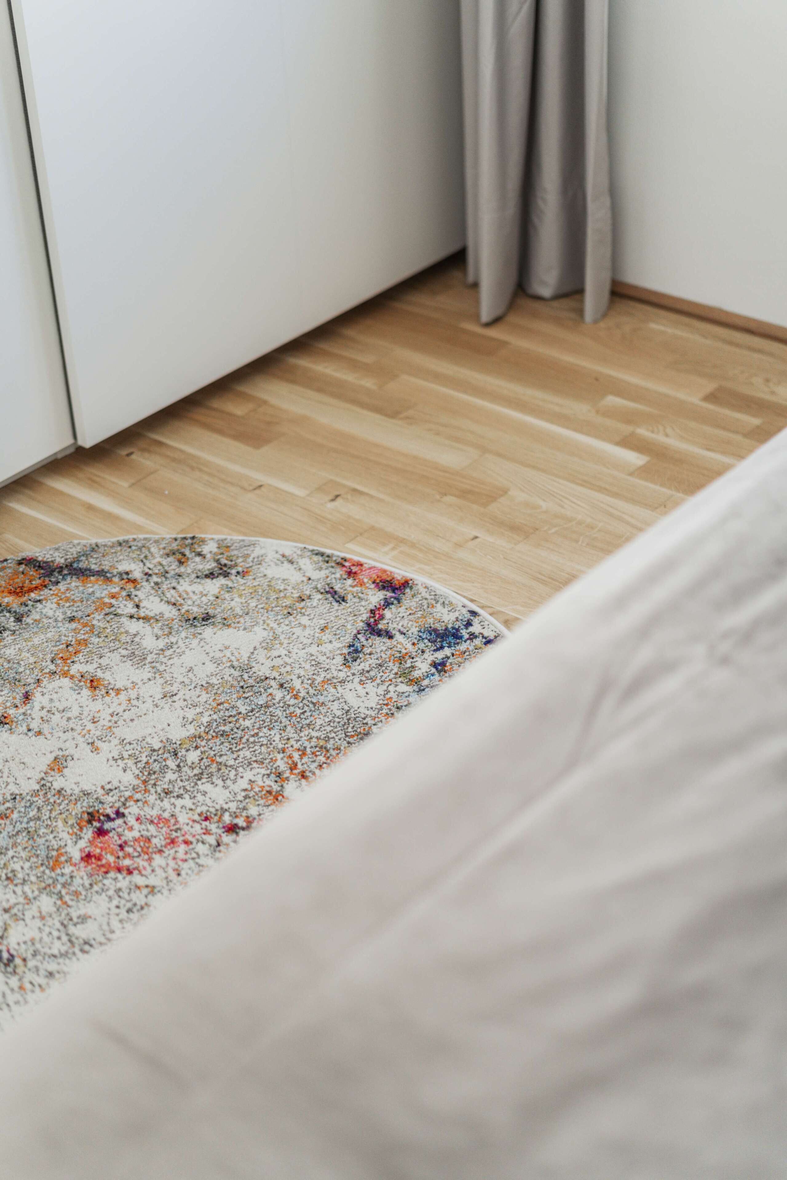 Una habitación con piso de madera, un tapete redondo con diseño multicolores y una cama blanca.