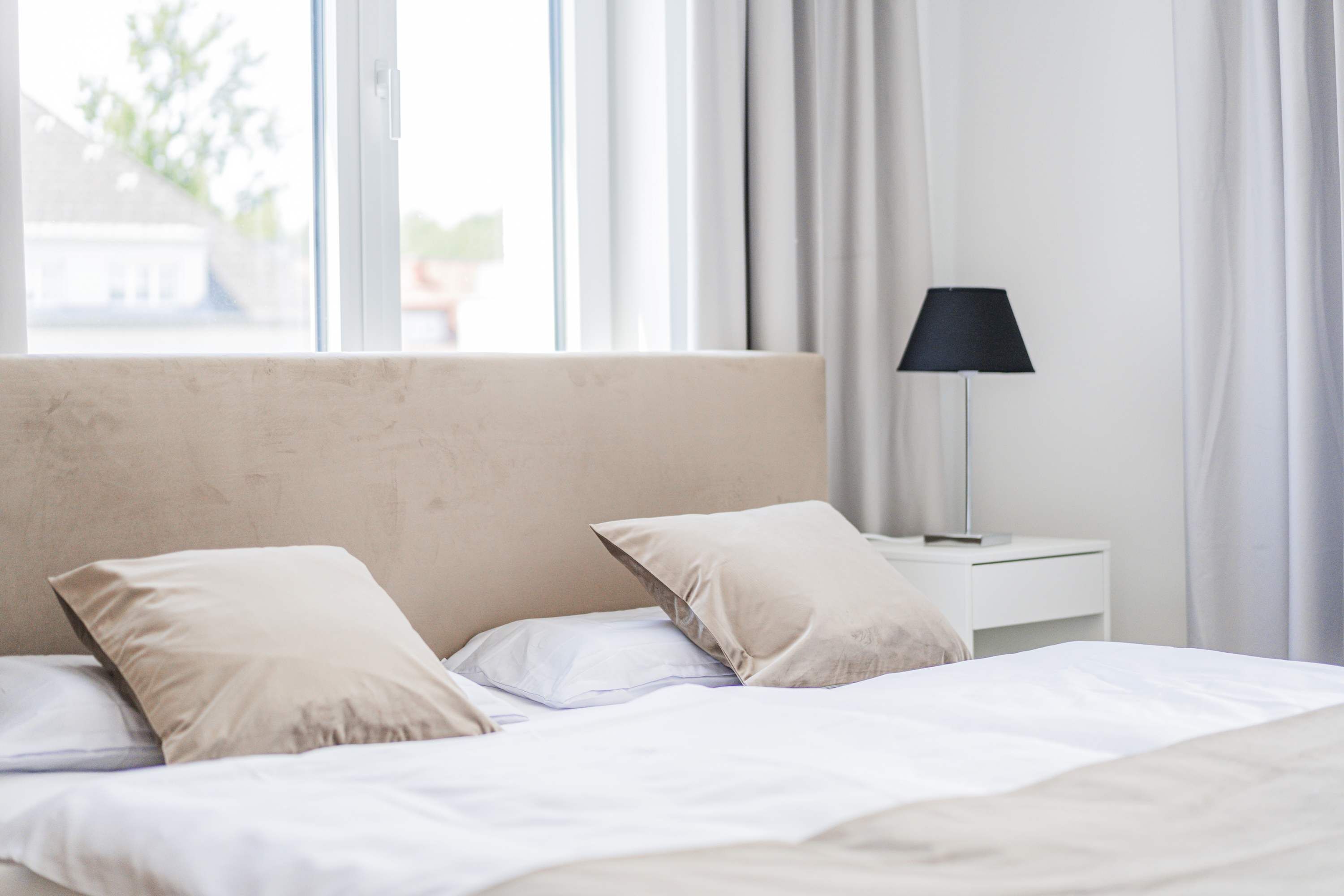 Un lit avec du linge de lit blanc et des oreillers beige, une lampe de chevet et une fenêtre avec des rideaux.