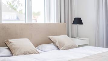 Un lit avec du linge de lit blanc et des oreillers beige, une lampe de chevet et une fenêtre avec des rideaux.