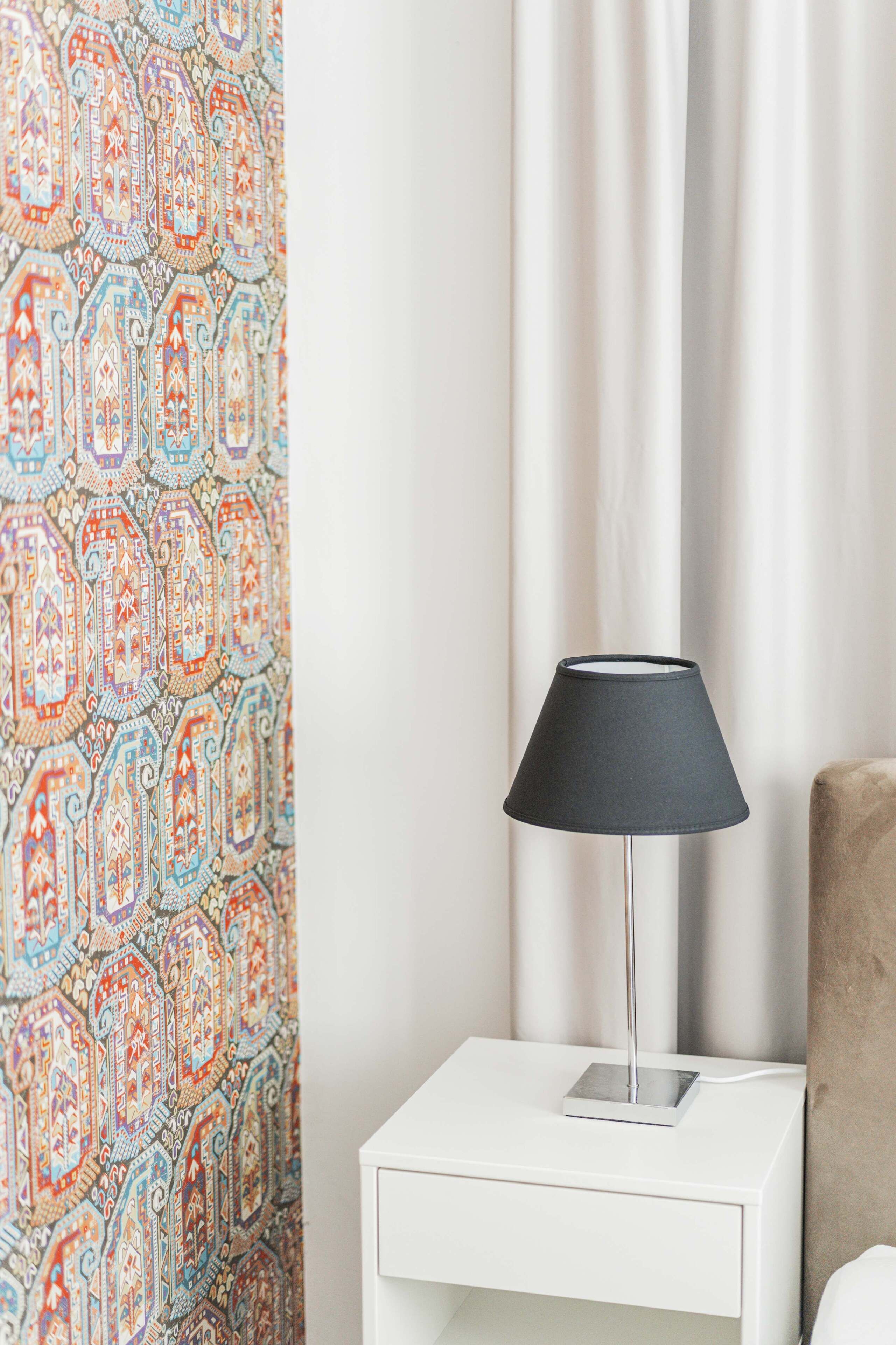 A white bedside table with a black lamp, a patterned curtain, and a plain wall.