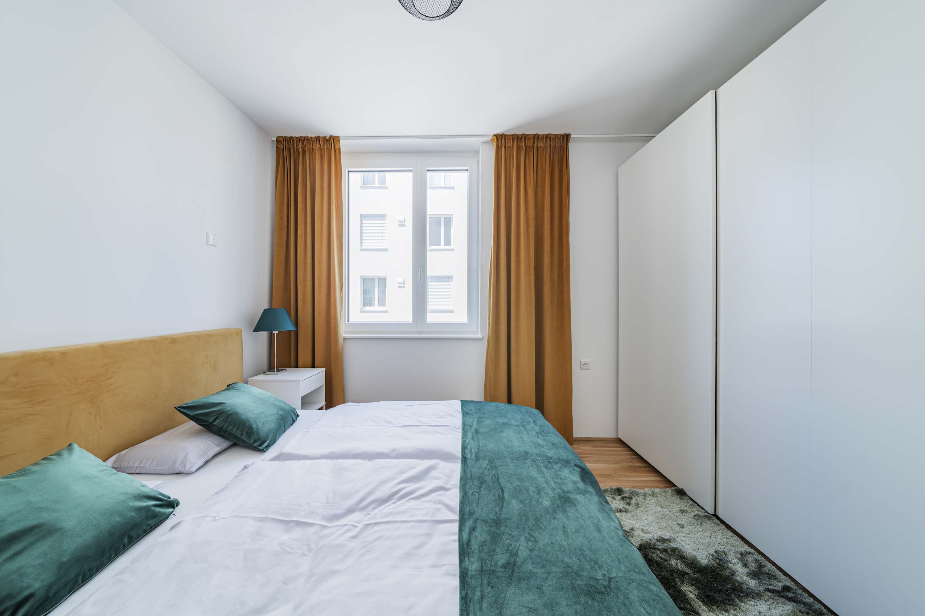 Een slaapkamer met een bed, nachtkastje, lamp en een raam met gordijnen.