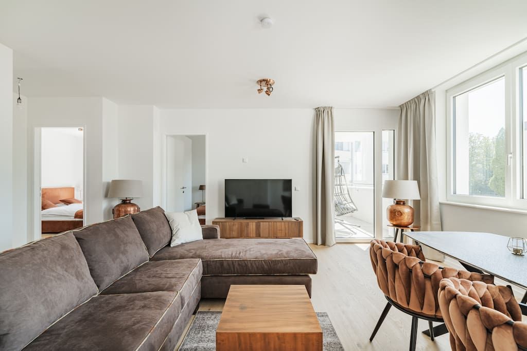 Ein modernes Wohnzimmer mit einem Ecksofa, einem Fernseher, einem Esstisch und Blick ins Freie.