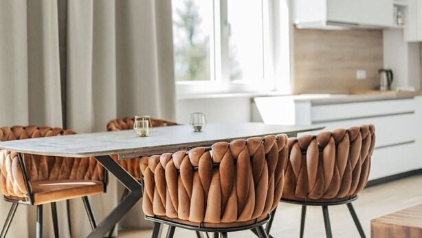 Et moderne spiseområde med et hvidt bord og polstrede brune stole, et køkken i baggrunden og udsigt til det fri gennem store vinduer.