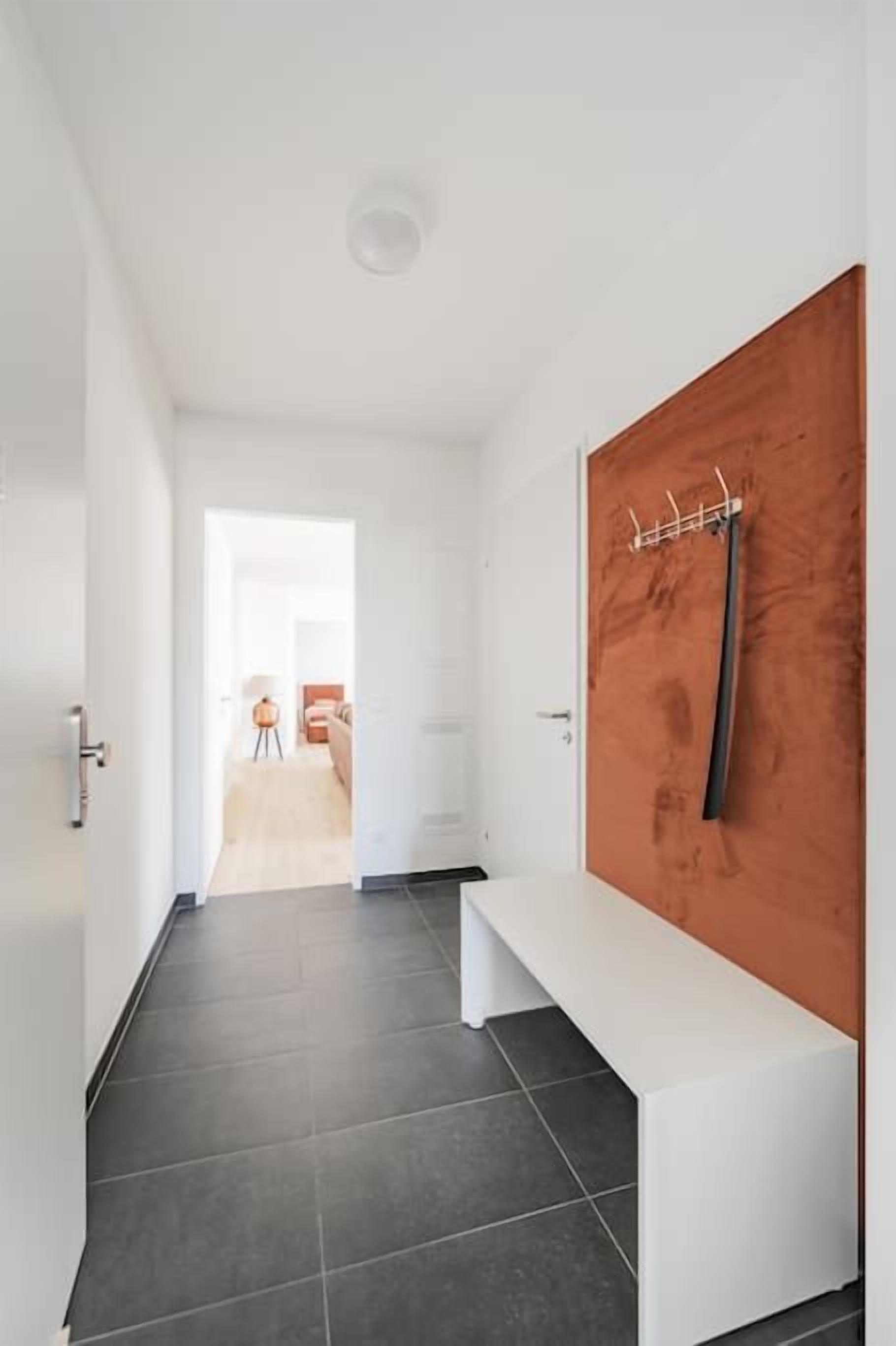 Un couloir avec un banc blanc, une porte en bois avec un porte-manteau, et une vue sur une autre pièce.