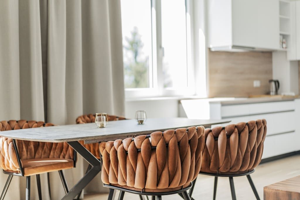 Un'area pranzo moderna con tavolo bianco e sedie imbottite in pelle marrone, una cucina sullo sfondo e vista all'esterno attraverso ampie finestre.