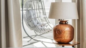 Et bord med en lampe og et glas, nær et vindue med udsigt til en hængestol og en bygning udenfor.