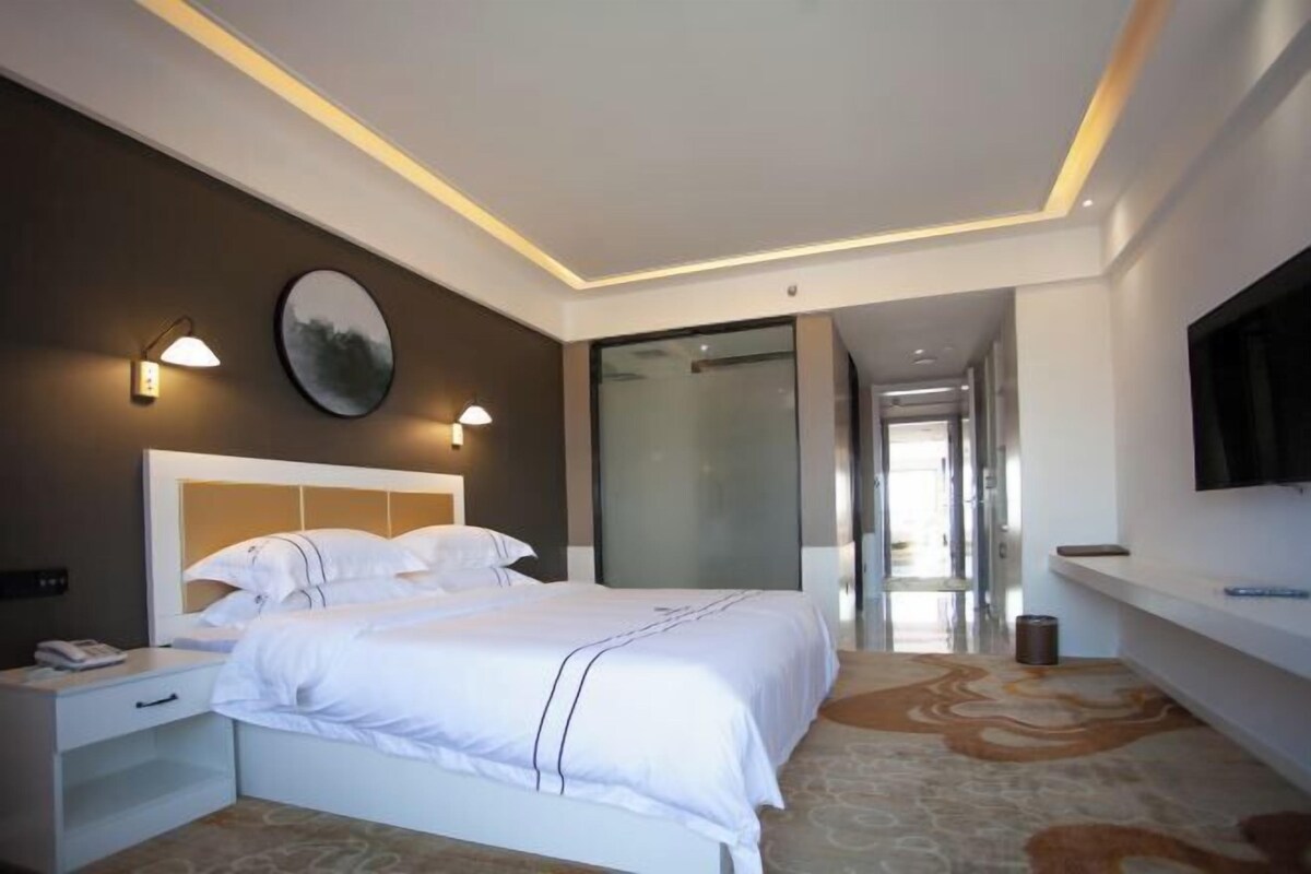 Ett modernt hotellrum med en stor säng, sängbord, en platt-TV och en glasdörr som leder ut till en hall.
