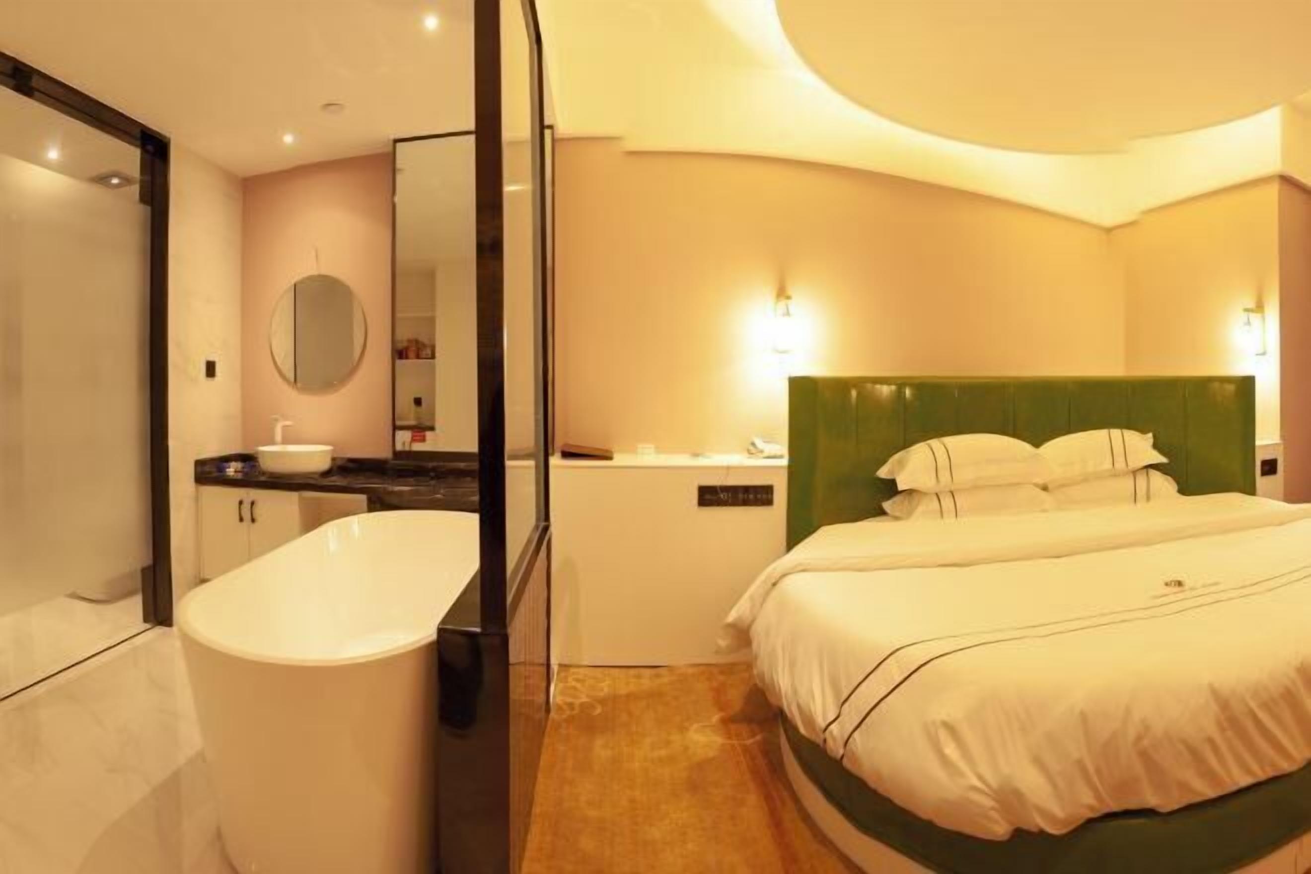 A hotel room with a large bathtub, a round mirror, a green headboard, and a white bed.