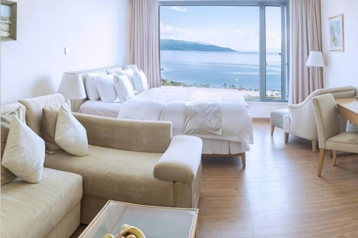 Een hotelkamer met een bed, een slaapbank, een fauteuil, een salontafel en een groot raam met uitzicht op zee.