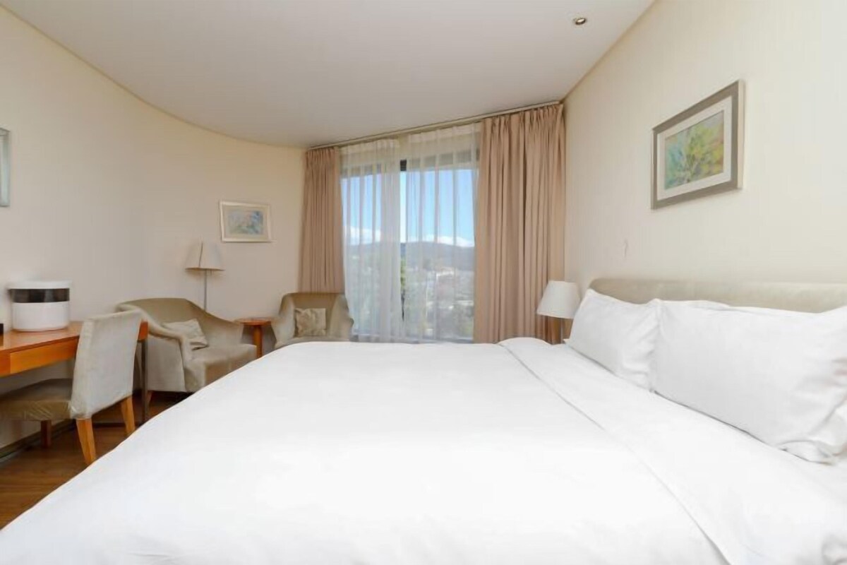 A hotel room with a large bed, a desk with a lamp, a chair, and a window with curtains.
