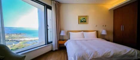 Habitación de hotel con un ventanal grande que ofrece vistas al mar y al paisaje, una cama con ropa blanca, una mesita de noche con lámparas y un armario de madera.
