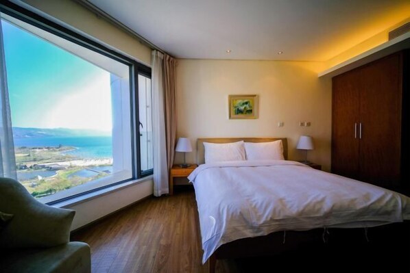 A hotel room with a large window offering a view of the sea and landscape, a bed with white linens, a nightstand with lamps, and a wooden wardrobe. - Huzhiyun Hotel (Chengjiang)